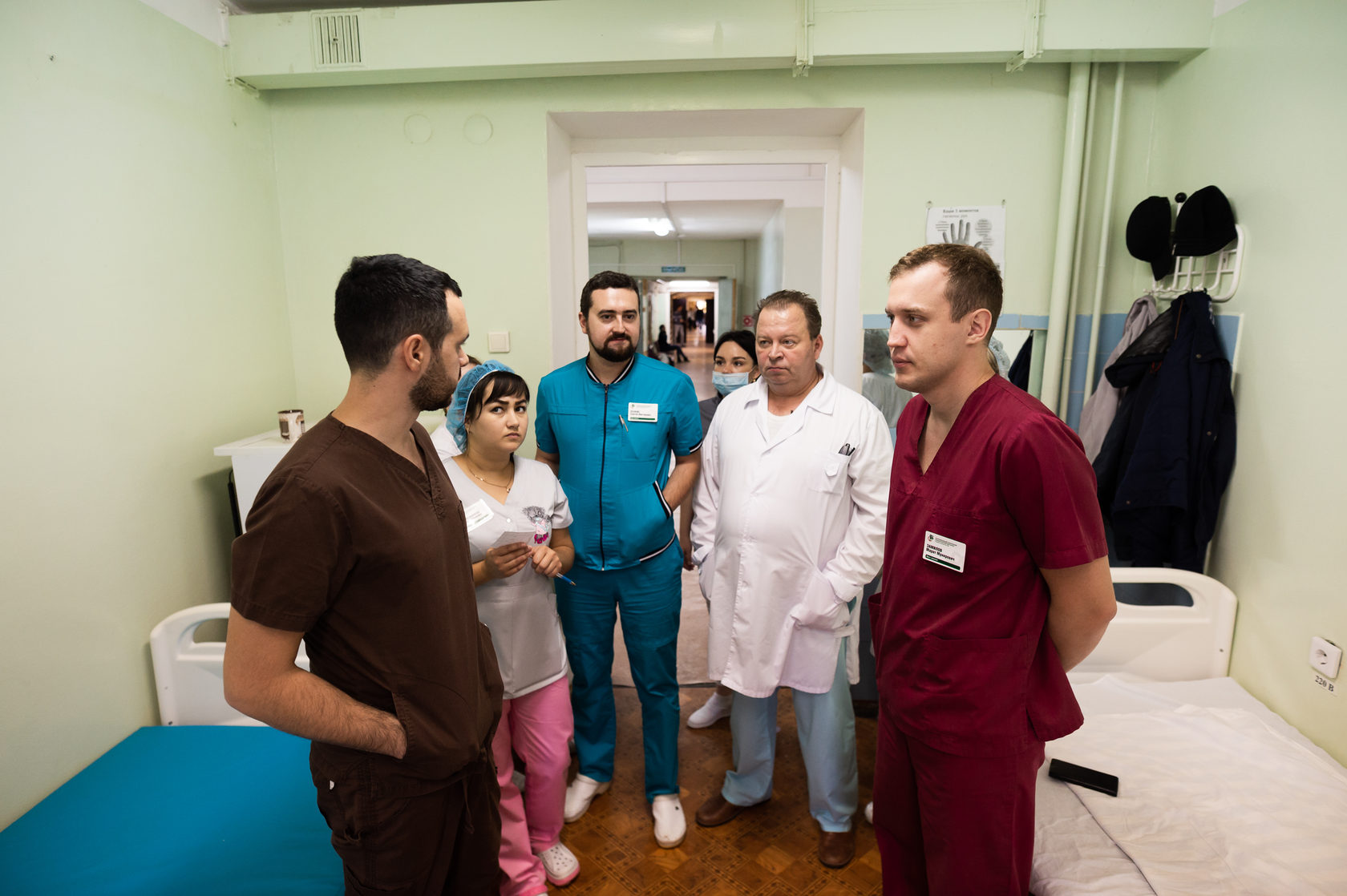 Группа людей стоит в больничной палате, между кроватей, слева направо: мужчина с короткими темными волосами и бородой, в темно-коричневом медицинский костюме; женщина с темными волосами, одетая в белый медицинский костюм, розовые брюки и голубую медицинскую шапочку; мужчина с короткими темными волосами и бородой, одетый в голубой медицинский костюм; мужчина с короткими темными волосами, одетый в белый медицинский халат; позади него стоит женщина в медицинской маске; мужчина с короткими темными волосами, одетый в бордовый медицинский костюм.
