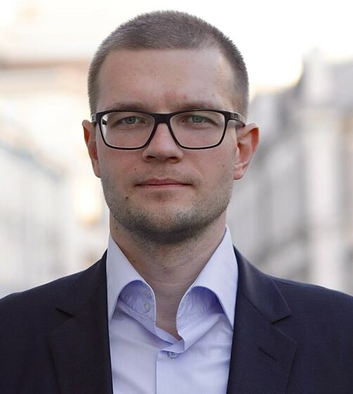 (Игорь Новиков) Мужчина с короткими темными волосами, короткой бородой и усами, в очках. Одет в голубую рубашку и темный пиджак.